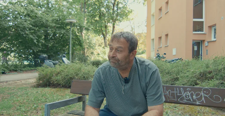 Alaattin T. sitzt auf einer Bank zwischen Bäumen, im Hintergrund ein Wohnhaus
