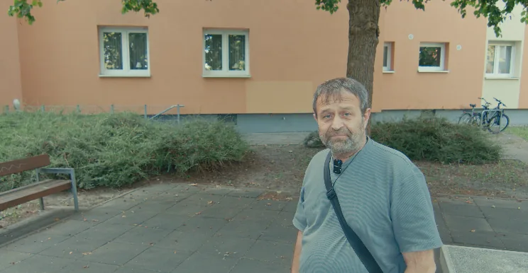 Alaattin T. steht auf einem gepflasterten Weg vor einem orangefarbenen Wohnhaus und blickt in die Kamera